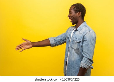 Side View Portrait Of Friendly Cheerful Man In Denim Casual Shirt With Rolled Up Sleeves Giving Hand To Handshake, Getting To Know New People, Meeting. Studio Shot Isolated On Yellow Background