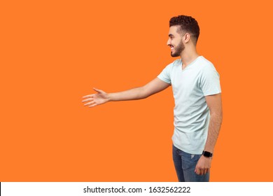 Side View Portrait Of Friendly Positive Man With Beard In White T-shirt Giving Hand To Handshake And Greeting, Empty Copy Space On Left For Text. Indoor Studio Shot Isolated On Orange Background