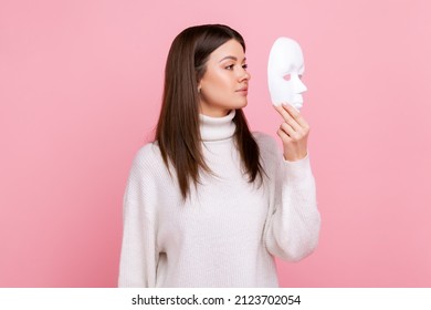 Side view portrait of girl holds white face mask, has mysterious expression, hiding his personality, wearing white casual style sweater. Indoor studio shot isolated on pink background.