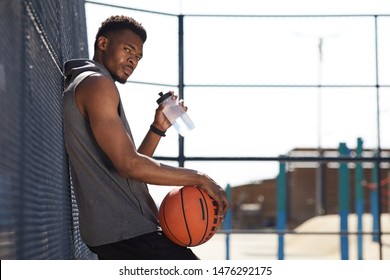 Side view portrait of handsome African-American man holding basketball ball looking at camera while drinking water in sports court outdoors, copy space - Powered by Shutterstock