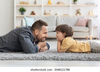 Side View Portrait Of Handsome Father And His Cute Little Son Looking At Each Other And Smiling, Lying On Floor At Home Interior, Free Space