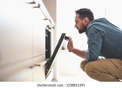 Side view portrait of handsome gentleman in denim shirt looking into oven while squatting on the floor