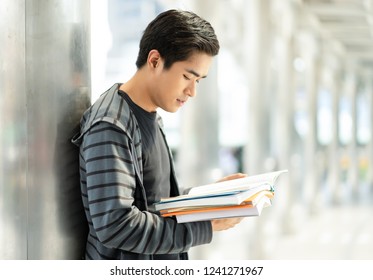 Side View Portrait Of A Handsome Male Student Standing Outside On Against A Wall Reading A Book