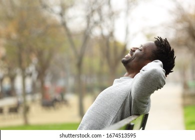 Side View Portrait Of A Happy Black Man Relaxing Sitting On A Bench In A Park