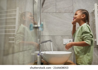 Side View Portrait Of Happy Black Kid With Braided Hair Brushing Teeth By Mirror In Bathroom, Copy Space