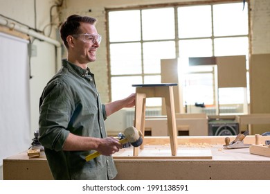 Side View Portrait Of Happy Carpenter Man In Safety Goggles Using Holding Hammer In Hands During Work, Having Handmade Stool Chair On Workbench, Smile. Copy Space.