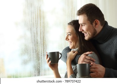 Side View Portrait Of A Happy Couple Holding Coffee Mugs Looking Outside Through A Window A Rainy Day Of Winter At Home