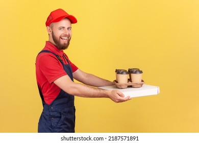 Side View Portrait Of Happy Deliveryman Standing With Pizza Box And Coffee In Disposable Cup And Looking Smiling At Camera, Fast Food Delivery Service. Indoor Studio Shot Isolated On Yellow Background