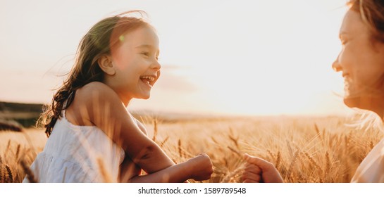 Side View Portrait Of A Happy Little Girl Laughing While Looking At Her Mother In A Field Of Wheat Against Sunset.