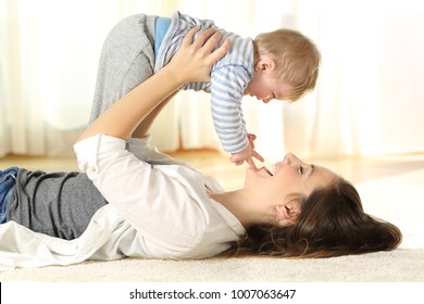 Side View Portrait Of A Happy Mother Lying On The Floor Raising Her Baby Son At Home