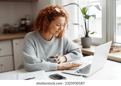 Side view portrait of happy redhead female web administrator sitting at kitchen table with copybook and smartphone, scrolling and typing on laptop, finishing her work while drinking coffee - Powered by Shutterstock