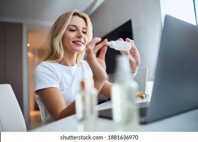 Side view portrait of happy smiling blonde woman holding plastic tube with cosmetic products in front of laptop camera in home apartment - Powered by Shutterstock