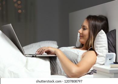 Side View Portrait Of A Happy Woman Typing In A Laptop On The Bed In The Night At Home