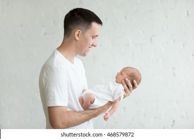 Side View Portrait Of Happy Young Father Holding His Newborn Sweet Adorable Baby Sleeping On His Arms. Both Dressed In White Unisex Clothes, Dad Looking At His Child Smiling.