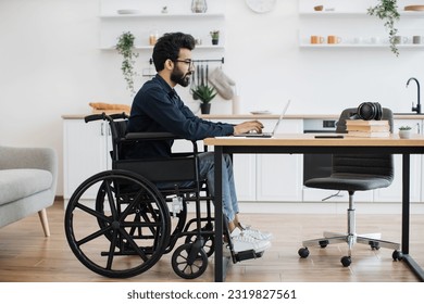 Side view portrait of indian male in wheelchair working at writing desk in open-plan kitchen of modern apartment. Young adult with disability starting working day with checking email account at home. - Powered by Shutterstock