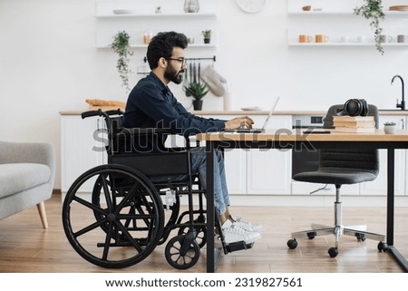 Side view portrait of indian male in wheelchair working at writing desk in open-plan kitchen of modern apartment. Young adult with disability starting working day with checking email account at home.