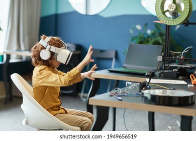 Стоковая фотография: Side view portrait of little boy wearing VR headset and reaching out while testing augmented technology in school laboratory, copy space