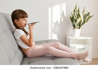 Side View Portrait Of Little Cute Dark Haired Female Child Wearing White Casual T Shirt Holding Phone, Sitting On Couch And Using Voice Assistant For Making Command.