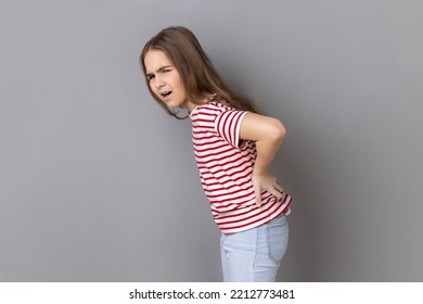 Side View Portrait Of Little Girl Wearing Striped T-shirt Screaming In Acute Pain And Holding Sore Back, Risk Of Kidney Stones Disease, Pinched Nerve. Indoor Studio Shot Isolated On Gray Background.