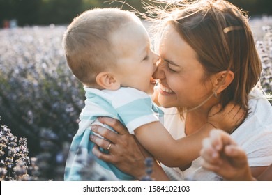 Side View Portrait Of A Lovely Kid Kissing Her Young Beautiful Mother Laughing In A Field Of Flower Against Sunset.