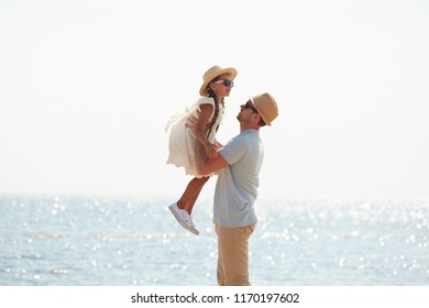 Side View Portrait Of Loving Father Playing With Little Girl By Sea, Picking Her Up And Smiling Happily, Copy Space