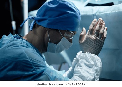 Side view portrait of male surgeon wearing mask and full protective clothing doing prayer ritual during surgery in operating room - Powered by Shutterstock