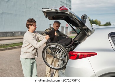 Side View Portrait Of Mature Couple Unloading Wheelchair Out Of Car Trunk In Parking Lot Outdoors, Copy Space