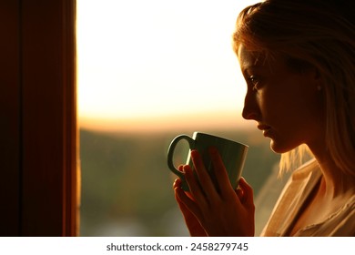 Side view portrait of a melancholic woman drinking coffee looking through a window at sunset - Powered by Shutterstock