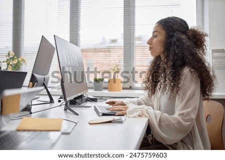 Side view portrait of Middle Eastern curly woman typing on keyboard at modern office against window. Concentrated female office worker designing website, or studying online
