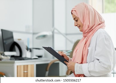 Side View Portrait Of Middle-Eastern Woman Wearing Hijab Working As Nurse In Medical Clinic And Writing On Clipboard, Copy Space