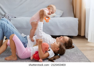 Side View Portrait Of Mom And Sweet Child Girls Lying And Playing On Floor, Mother Holding Infant Baby Ina Hands, Family Expressing Positive Emotions And Happiness.