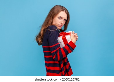 Side View Portrait Of Offended Woman Wearing Striped Sweater, Embracing Wrapped Gift Box, Being Greedy, Does Not Want To Give Present, Looking At Camera. Indoor Studio Shot Isolated On Blue Background