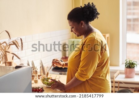 Side view portrait of overweight black woman cooking healthy meal in kitchen lit by sunlight, copy space