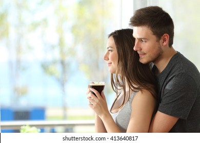 Side View Portrait Of A Pensive Couple Or Marriage Hugging And Looking Outdoors Through A Window Of An Hotel Room Or Home With The Sea In The Background