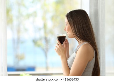 Side view portrait of a pensive girl drinking coffee and looking outdoors through a window of an hotel room or home with the sea in the background