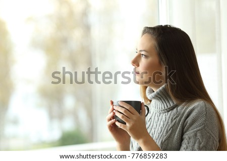 Side view portrait of a pensive serious woman looking through a window at home in a sad day