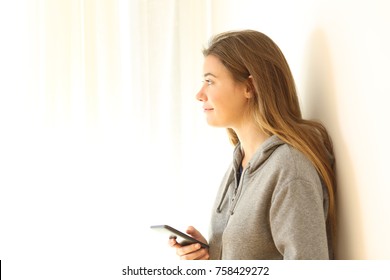 Side View Portrait Of A Pensive Teen Holding A Smart Phone Looking Through A Window Isolated On White At Side