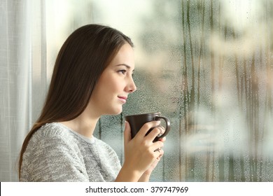 Side View Portrait Of A Pensive Woman Looking Away Through A Wet Window In A Rainy Day At Home