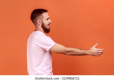 Side View Portrait Of Positive Friendly Handsome Bearded Man Holding Out His Hand To Side, Giving Handshake, Wearing Pink T-shirt. Indoor Studio Shot Isolated On Orange Background.