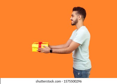 Side view portrait of positive generous brunette man with beard in white t-shirt smiling and giving gift box, sharing holiday present, charity concept. indoor studio shot isolated on orange background
