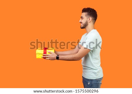 Side view portrait of positive generous brunette man with beard in white t-shirt smiling and giving gift box, sharing holiday present, charity concept. indoor studio shot isolated on orange background