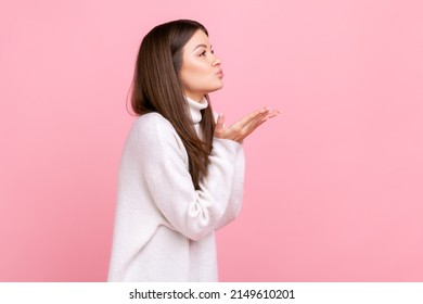 Side view portrait of pretty falling in love female sending air kissing, expressing romance, flirts, wearing white casual style sweater. Indoor studio shot isolated on pink background.