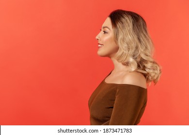 Side view portrait of pretty girl with blonde hair in brown blouse, she looks to the side, smiles, express happiness, energy and friendliness. Indoor studio shot isolated on red background - Shutterstock ID 1873471783