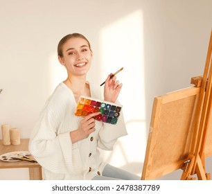 Side view portrait of a pretty woman painter drawing with watercolor palette near easel. FEmale artist painter on canvas in creative studio. - Powered by Shutterstock