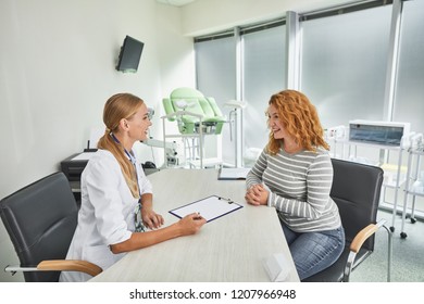 Side view portrait of red-haired woman sitting across the table from gynecologist. They looking at each other and smiling - Shutterstock ID 1207966948