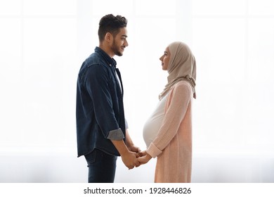 Side View Portrait Of Romantic Pregnant Muslim Couple Holding Hands At Home, Young Arabic Spouses Expecting For A Baby, Standing Together Near Window, Looking At Each Other With Love, Copy Space