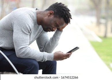 Side View Portrait Of A Sad Black Man Complaining Checking Mobile Phone Sitting On A Bench In A Park