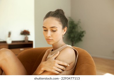 Side view portrait of sad melancholic beautiful caucasian girl coping with grief, looking down touching her shoulder sitting on couch in her living-room. Negative human emotions and feelings
