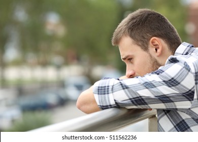 Side view portrait of a sad single man looking down from a balcony of a house  with an urban background