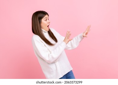 Side view portrait of scared female with raised arms, sees something awful, afraids, stop gesture, wearing white casual style sweater. Indoor studio shot isolated on pink background.
